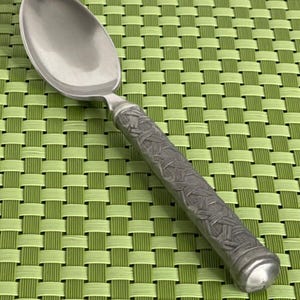 May include: A silver spoon with an ornate, textured handle. The spoon is resting on a green woven placemat.