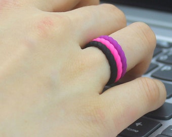May include: A hand wearing three silicone rings stacked on top of each other. The rings are black, pink, and purple.