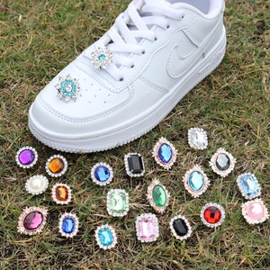May include: A white trainer with various colourful rhinestone embellishments. The rhinestones are in different shapes and sizes, including round, square, and oval. The rhinestones are arranged in a random pattern on the trainer.