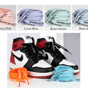 May include: A pair of black and red high-top sneakers with white laces. The sneakers are sitting on a white surface with four different colored shoelaces in the background. The shoelaces are labeled "SHOELACES" in black text. The colors of the shoelaces are cherry pink, coral blue, bean green, and blue grey.