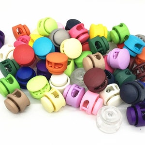 May include: A collection of colorful plastic toggle clasps, also known as cord locks, in various shapes and sizes. The clasps are used for adjusting the length of cords, drawstrings, and straps.