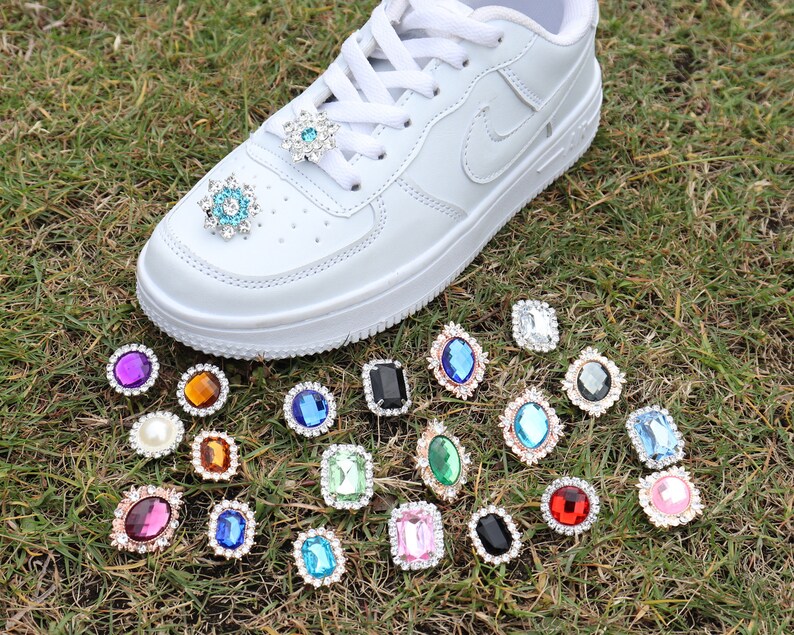 May include: A white trainer with various colourful rhinestone embellishments. The rhinestones are in different shapes and sizes, including round, square, and oval. The rhinestones are arranged in a random pattern on the trainer.