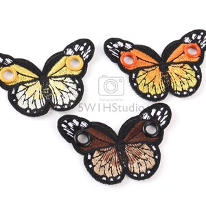 May include: Three embroidered butterfly shoe charms with black outlines and colorful wings. The butterflies have yellow, orange, and brown wings with black and white accents. Each butterfly has two eyelets for attaching to shoes.