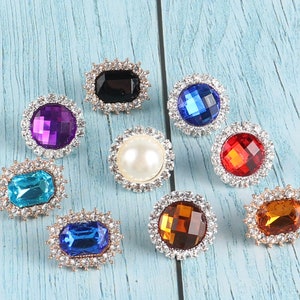 May include: Assortment of eight decorative rhinestone embellishments in various colours and shapes, including round, square, and rectangular. The rhinestones are set in silver-toned metal settings.