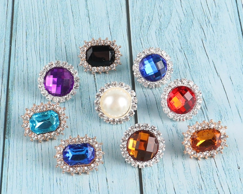 May include: Assortment of eight decorative rhinestone embellishments in various colours and shapes, including round, square, and rectangular. The rhinestones are set in silver-toned metal settings.