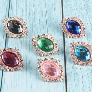 May include: Six oval-shaped, jeweled brooches with gold-toned settings. The brooches feature various colours, including black, green, blue, turquoise, pink, and burgundy.