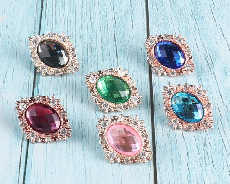 May include: Six oval-shaped, jeweled brooches with gold-toned settings. The brooches feature various colours, including black, green, blue, turquoise, pink, and burgundy.