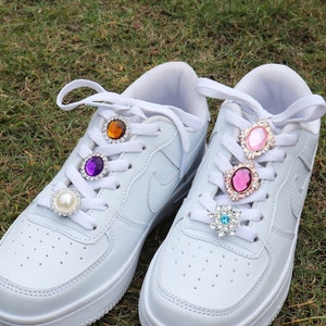 May include: A pair of white trainers with colourful jeweled shoe charms on the laces. The charms are purple, orange, pink, and blue.