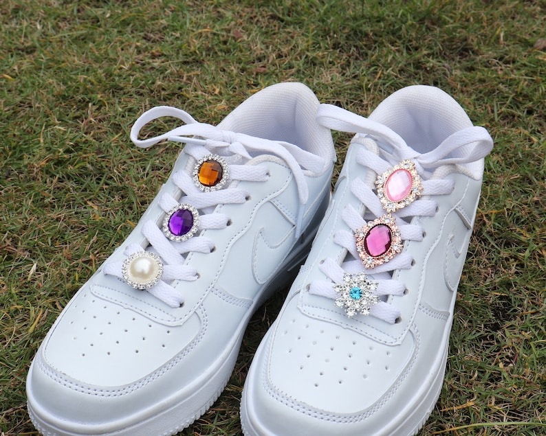 May include: A pair of white trainers with colourful jeweled shoe charms on the laces. The charms are purple, orange, pink, and blue.