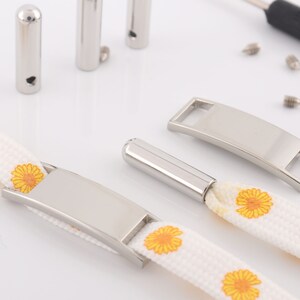 May include: A white canvas strap with yellow daisy print and two silver metal buckles. One buckle is a rectangular tag with a slot for personalization. The other buckle is a curved bar with a hole for attaching a key ring or other item.