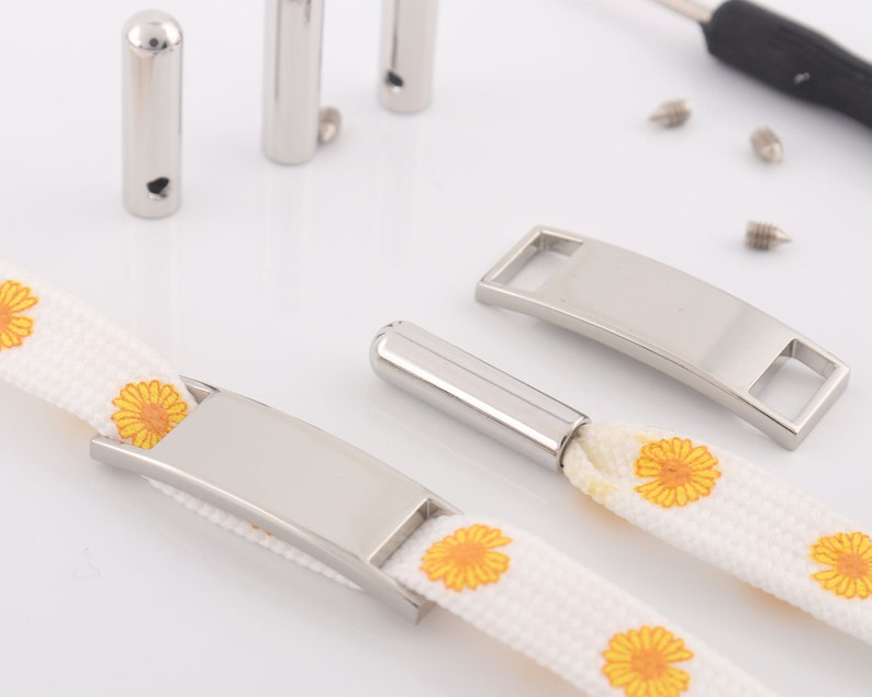 May include: A white canvas strap with yellow daisy print and two silver metal buckles. One buckle is a rectangular tag with a slot for personalization. The other buckle is a curved bar with a hole for attaching a key ring or other item.