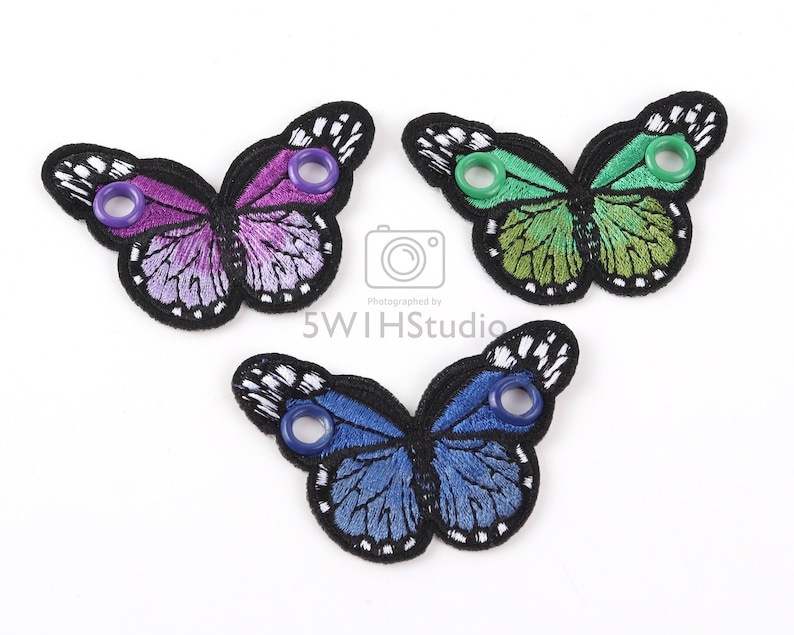 May include: Three embroidered butterfly shoe charms in purple, green, and blue with black outlines and white accents. Each charm has two eyelets.