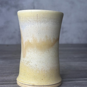 May include: A handmade ceramic tumbler with a flared base and a slightly curved body. The exterior features a speckled, layered design with shades of cream, yellow, and tan. The rim is smooth, and the overall aesthetic is rustic and organic.