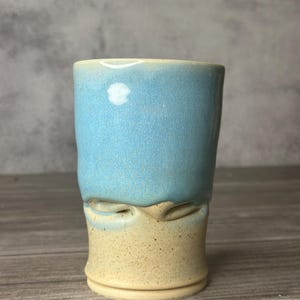 May include: A handmade ceramic tumbler with a two-tone design. The top half is a light blue, while the bottom half is a speckled tan. The tumbler has a unique, textured appearance with a slightly irregular shape, suggesting it's handcrafted.