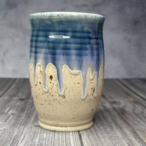 May include: A ceramic tumbler featuring a glossy blue glaze on the upper portion that drips down over a speckled cream-colored base. The surface displays a visible crackle finish within the blue glaze, creating a textured appearance on the vessel.