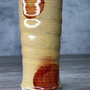 May include: A handmade ceramic tumbler with a beige body and a reddish-brown glaze. The cylindrical cup has a textured, ridged design and a white base. The glaze creates a unique, organic pattern, adding to its rustic charm.