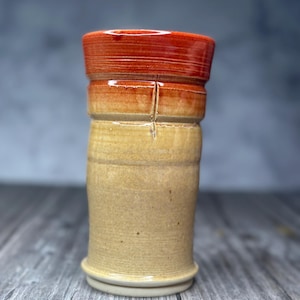 May include: A cylindrical ceramic tumbler featuring a two-tone glaze. The upper section is a vibrant, glossy orange-red, while the lower portion transitions into a matte, sandy beige finish. A vertical incised line runs through the center.