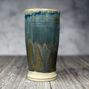 May include: A tall, cylindrical ceramic tumbler featuring a reactive glaze with a blend of deep blue, teal, and earthy olive green tones. The base is unglazed, showing a smooth, light beige clay finish with a subtle raised ring detail at the bottom.