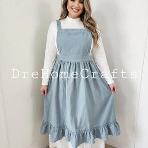 May include: A light blue apron dress with a ruffled hem. The dress has a square neckline and wide straps. The text "DreHomeCrafts" is printed on the dress. The dress is worn over a white long-sleeved top.