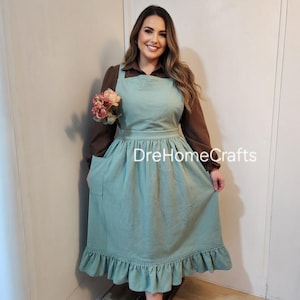 Puede incluir: Un delantal verde azulado claro con dobladillo con volantes y un bolsillo grande. El delantal se usa sobre una camisa marrón de manga larga. El delantal tiene un escote cuadrado y una cintura entallada. El texto "DreHomeCrafts" es visible.