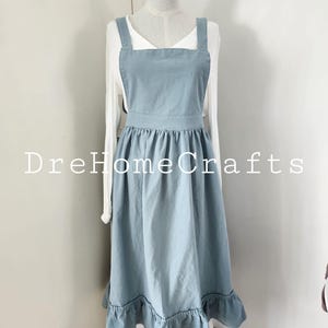May include: A light blue apron with a ruffled hem and square neckline is displayed on a mannequin. The apron has wide shoulder straps and a fitted waist. The text "DreHomeCrafts" is printed on the apron. The apron is worn over a white long-sleeved shirt.