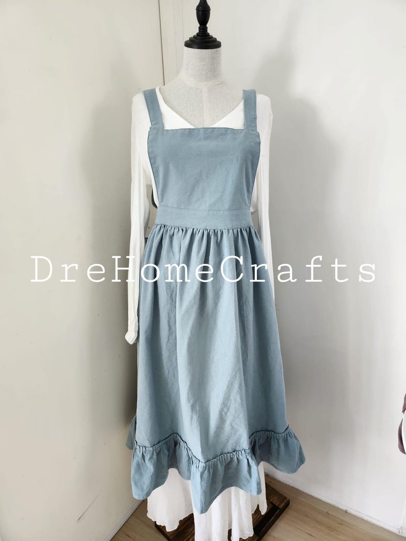 May include: A light blue apron with a ruffled hem and square neckline is displayed on a mannequin. The apron has wide shoulder straps and a fitted waist. The text "DreHomeCrafts" is printed on the apron. The apron is worn over a white long-sleeved shirt.