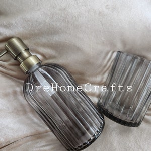 May include: A set of two gray glass bathroom accessories with a ribbed design. The set includes a soap dispenser with a gold pump and a tumbler.