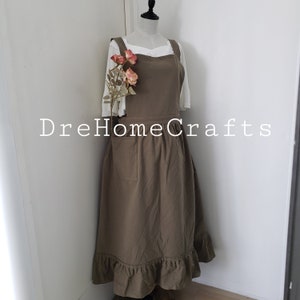May include: A brown apron with a ruffled hem and a large front pocket is displayed on a mannequin. The apron has a square neckline and wide straps. The text "DreHomeCrafts" is printed on the apron. A white blouse and pink flowers are also visible.
