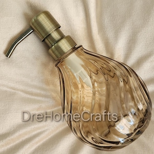 May include: A soap dispenser with a teardrop-shaped, amber-tinted glass body featuring vertical ribbed textures. The top includes a metallic, antique brass-finished pump mechanism. The text DreHomeCrafts is printed in white on the glass surface.