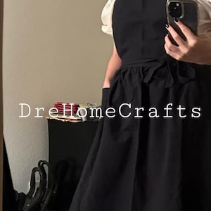 May include: Black apron with a ruffled hem and a white blouse underneath. The apron has a tie at the waist and pockets. The text "DreHomeCrafts" is visible on the image.
