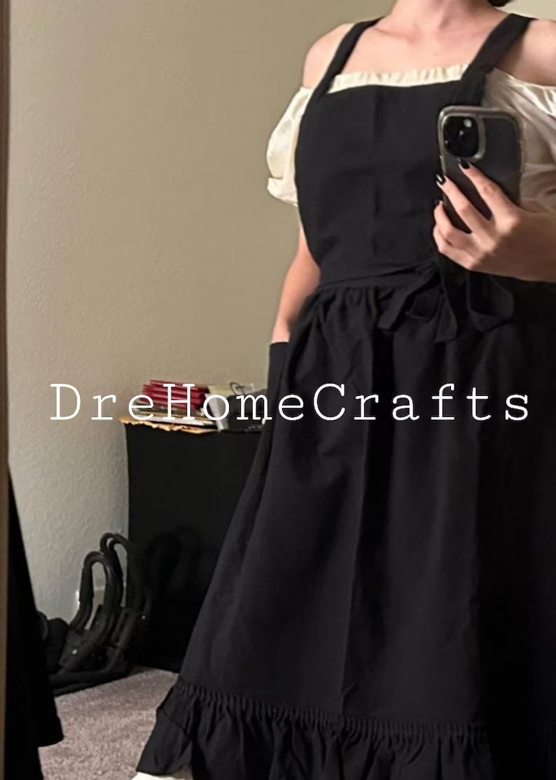 May include: Black apron with a ruffled hem and a white blouse underneath. The apron has a tie at the waist and pockets. The text "DreHomeCrafts" is visible on the image.