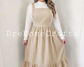 Peut inclure: Une robe tablier beige avec un ourlet à volants et une encolure carrée. Le tablier a une poche sur le côté droit et est porté sur un haut blanc à manches longues. Le texte "DreHomeCrafts" est imprimé sur le tablier.