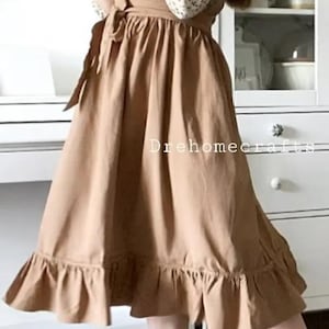 May include: A tan apron dress with a ruffled hem. The dress is tied at the waist and worn over a floral print blouse. The image includes the text "Drehomecrafts". The person is holding a bouquet of flowers.