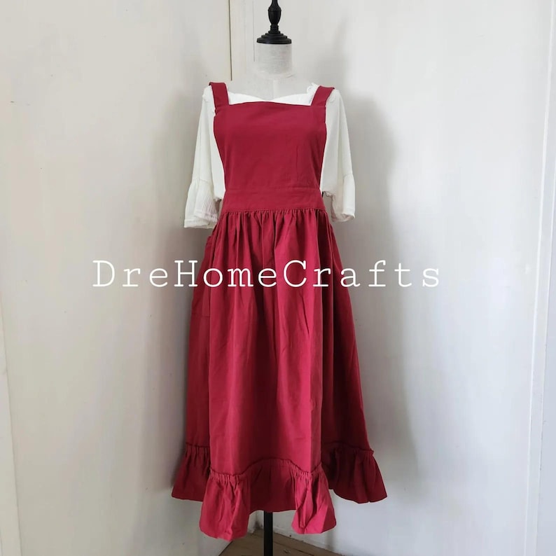 May include: A red apron dress with a white blouse underneath, displayed on a mannequin. The dress has a square neckline, wide straps, and a ruffled hem. The text "DreHomeCrafts" is visible.
