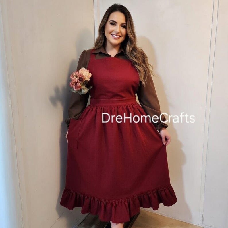 May include: A burgundy apron dress with a ruffled hem and pockets. The dress is worn over a brown long-sleeved shirt. The text "DreHomeCrafts" is visible on the dress.