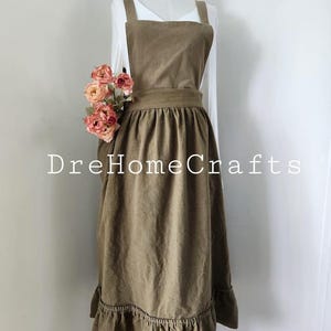 May include: A brown apron with a ruffled hem and a square neckline. The apron has a wide waistband and shoulder straps. The text "DreHomeCrafts" is printed on the apron. A bouquet of pink flowers is placed on the left side of the apron.