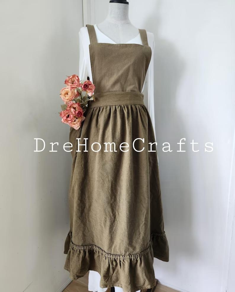 May include: A brown apron with a ruffled hem and a square neckline. The apron has a wide waistband and shoulder straps. The text "DreHomeCrafts" is printed on the apron. A bouquet of pink flowers is placed on the left side of the apron.
