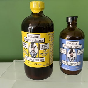 May include: Two glass bottles with yellow and black labels. The larger bottle is labeled "DOCKTOR FIRWAT AISYA" and "BLACK GARLIC FIRE CIDER". The smaller bottle is labeled "DOCKTOR FIRWAT AISYA" and "ORGANIC ELDERBERRY SYRUP".