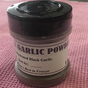 May include: A small glass jar with a black lid filled with dehydrated black garlic powder. The label reads "Garlic Powder Dehydrated Black Garlic Best by: Place - Best in Freezer".