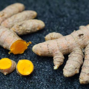 May include: Close-up of fresh turmeric roots, showcasing their earthy brown exterior and vibrant orange interior. Several whole and sliced pieces are arranged on a dark, textured surface, highlighting the spice's natural form.