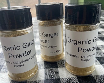May include: Three glass jars filled with organic ginger powder. The jars have black lids and white labels with the text "Organic Ginger Powder" and "Ingredients: Dehydrated Ginger". The jars are sitting on a black and white checkered tablecloth.