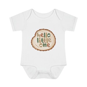 May include: White baby onesie with a woodgrain circle design featuring the text "hello little one" in green, brown, and orange.