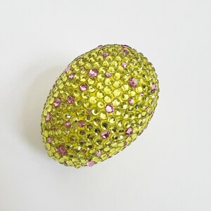 May include: A decorative egg covered in yellow and pink rhinestones. The egg-shaped object is covered in small, sparkling rhinestones in shades of yellow and pink. The egg is set against a plain white background.