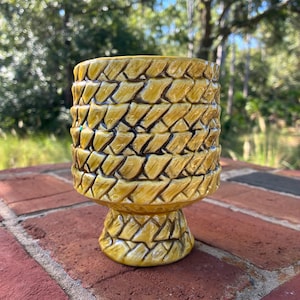 May include: A mustard yellow ceramic planter with a raised, woven basket-weave texture across the exterior. The vessel features a cylindrical body that tapers down to a pedestal base, with dark brown glaze accents highlighting the woven pattern.