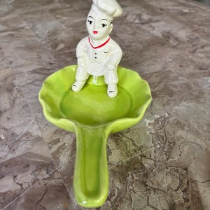 May include: A ceramic spoon rest shaped like a bright green lettuce leaf with a handle. A small, white-clad chef figurine with a tall hat, red neck trim, and black buttons sits upright in the center of the leaf, facing forward with painted features.