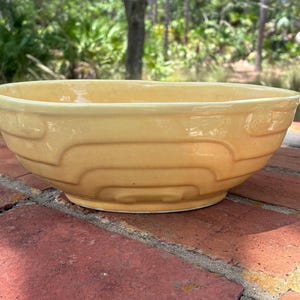 May include: A pale yellow ceramic mixing bowl featuring a glossy finish and a series of concentric, stepped, rectangular embossed patterns encircling the exterior side. The bowl has a wide, open top and a smooth, rounded base design.