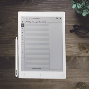 Pu&ograve; includere: Un e-reader bianco con uno schermo grigio che mostra un elenco di cose da fare intitolato "Priority / to do for the week of". L'elenco ha caselle di controllo e righe per scrivere. L'e-reader ha una penna stilo nera e grigia accanto.