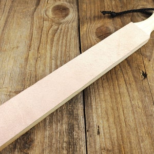 Puede incluir: Un asentador de navaja recta con una superficie de cuero de color claro y un mango de madera. El asentador es rectangular, con una superficie de cuero de color beige claro. El mango es de madera clara y tiene un agujero para colgar.