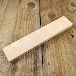 Puede incluir: Un cuero rectangular de color beige claro para afilar cuchillos. El cuero está montado sobre una base de madera. La veta de la madera es visible en el fondo.