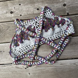 Puede incluir: Top de crochet blanco hecho a mano con un dise&ntilde;o floral en el centro. El top presenta un cuello halter y correas anudadas, con un patr&oacute;n colorido y multicolor en el dise&ntilde;o floral. El crochet es abierto y aireado.